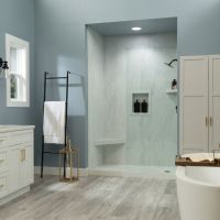 Coastal-inspired bathroom featuring Arctic Vein marble-look shower wall panels, soft blue walls, a white shaker vanity with gold hardware, light gray wood-look flooring, a frameless glass shower, and a freestanding soaking tub.