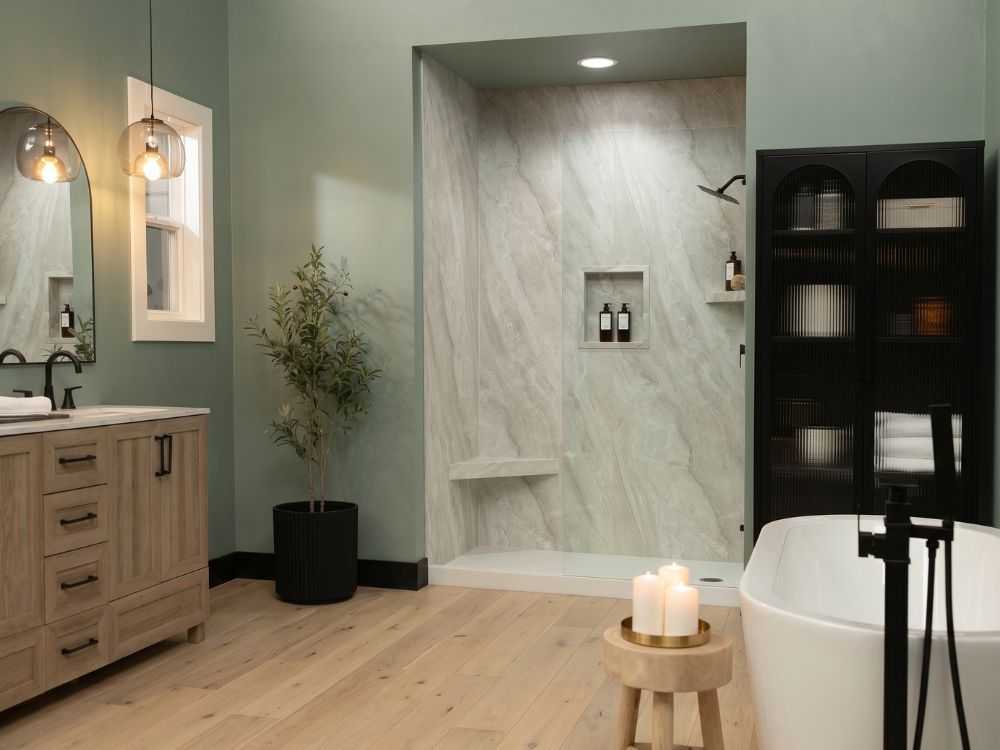 Contemporary bathroom featuring Crema Marfil marble-look shower wall panels, sage green walls, a light oak vanity, matte black fixtures, light wood flooring, and a freestanding soaking tub beside a spacious walk-in shower.