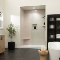 Modern bathroom featuring Tuscany Sand shower wall panels in a 36x24 tile pattern with white joints, light wood vanity cabinetry, matte black fixtures, dark gray floor tile, a freestanding soaking tub, and a spacious walk-in shower with built-in storage niches.