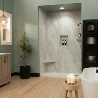 Contemporary bathroom featuring Crema Marfil marble-look shower wall panels, sage green walls, a light oak vanity, matte black fixtures, light wood flooring, and a freestanding soaking tub beside a spacious walk-in shower.