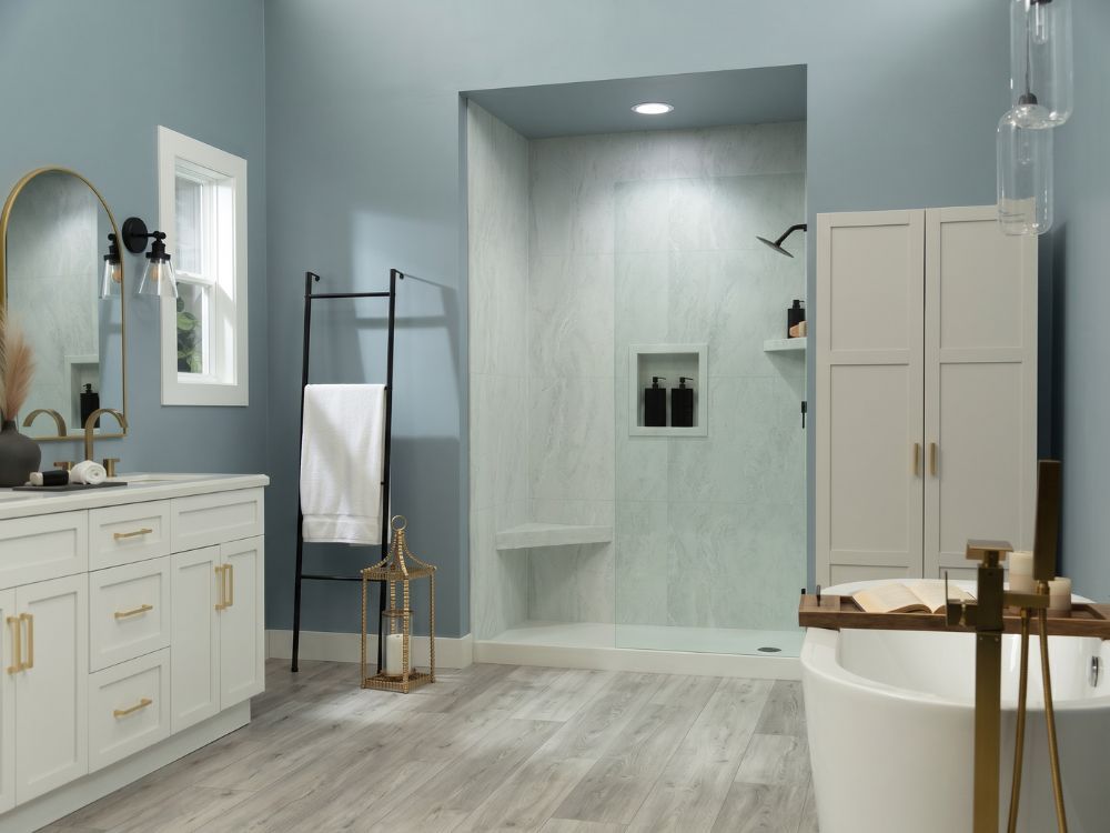 Coastal-inspired bathroom featuring Arctic Vein marble-look shower wall panels, soft blue walls, a white shaker vanity with gold hardware, light gray wood-look flooring, a frameless glass shower, and a freestanding soaking tub.